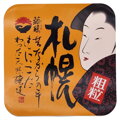 Detailný pohľad na oranžový vrchnák plastového obalu hrubo mletej Miso pasty Shih-Chuan. Obsahuje tradičnú japonskú kresbu tváre, výraznú čiernu kaligrafiu a červenú pečať.